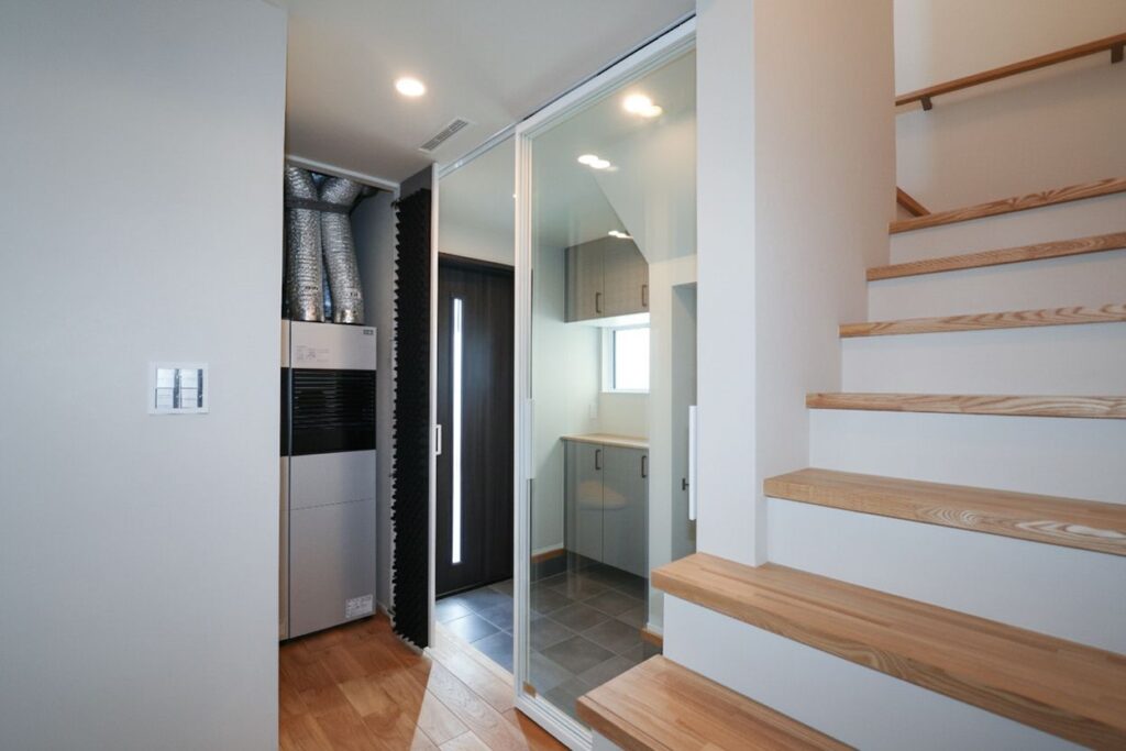 自然光が差し込む玄関ホール。手前には木目調の階段があり、奥には全館空調ユニットが見える。ガラスの引き戸で仕切られた土間には、グレーのキャビネットと、縦長の採光窓のある黒いドアが配置された、機能的で広々とした住宅のインテリア。