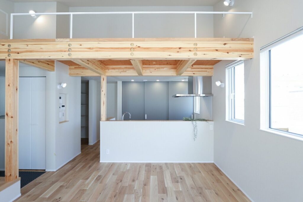 吹き抜け空間を活用したロフト構造のLDK。白壁に映える木製の太い梁と柱、対面式のキッチン、そして自然光が差し込む大きな窓がある、明るく開放感のある注文住宅の室内写真。
