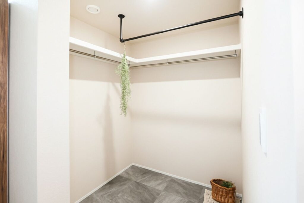 L字型の枕棚とハンガーパイプ、天井に設置された黒いアイアンバーが特徴的なウォークインクローゼット。グレーのフロアタイルと白い壁が調和した、収納力と室内物干し機能を兼ね備えた空間です。