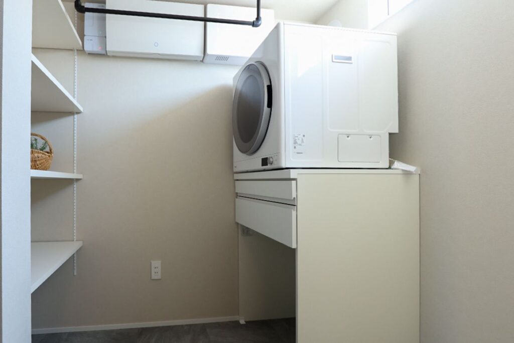 白を基調とした機能的なランドリールーム。引き出し収納付きの専用台に設置された衣類乾燥機、天井の黒いアイアンバー、左手の可動棚が備わり、洗濯から室内干し、収納までスムーズに行える家事動線の良い空間です。