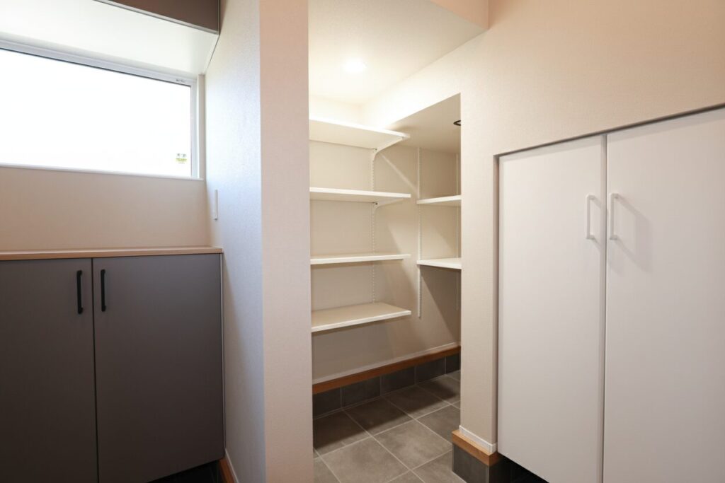 自然光が入る窓付きの明るい土間収納スペース。白い壁に設置された高さ調節可能なオープンシェルフと、グレーおよびホワイトの扉付きキャビネットを組み合わせた、機能的で広々とした住宅の玄関インテリア。