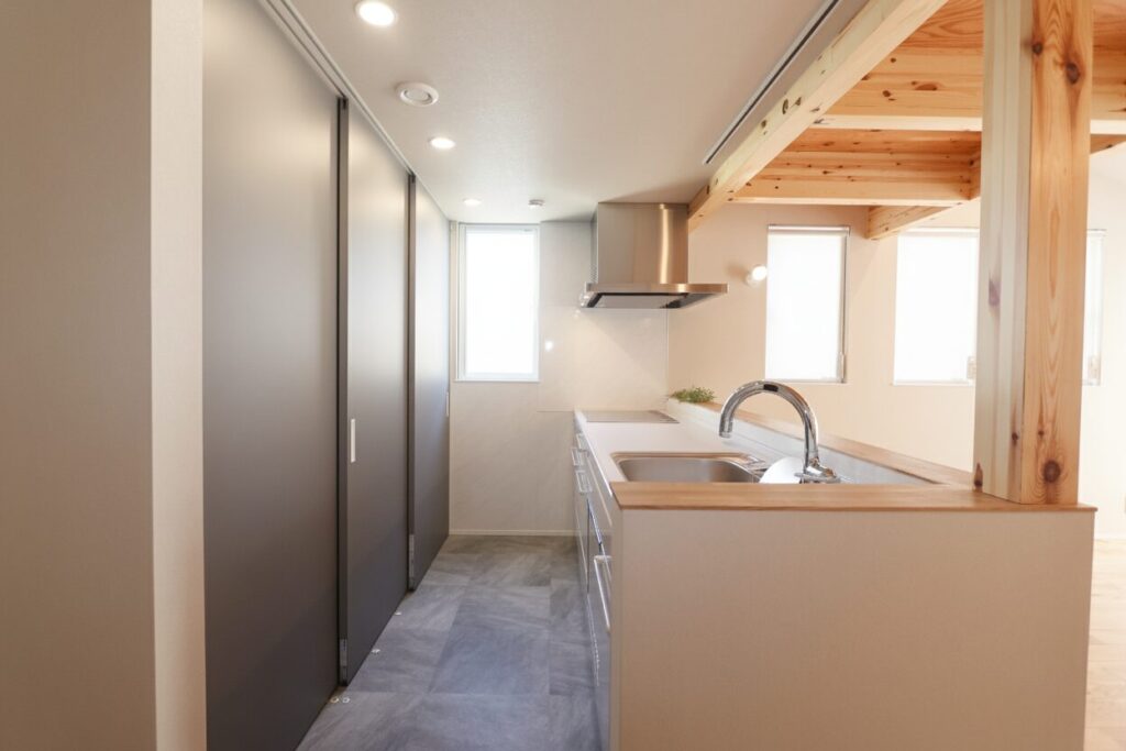 木の梁と柱がアクセントになったナチュラルモダンな対面式キッチン。グレーの引き戸式背面収納とフロアタイルが落ち着いた空間を演出しています。