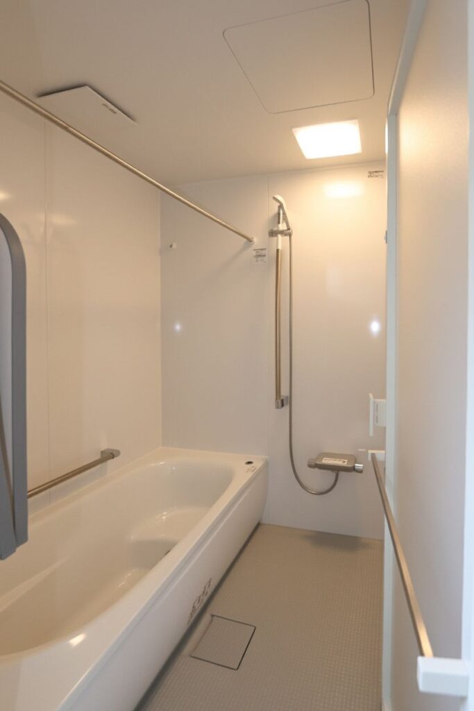 白を基調とした清潔感あふれるシステムバス。浴槽の横や出入り口に設置された手すり、高さを調節できるスライドバー付きシャワー、浴室干し用のランドリーパイプを備えた、明るく安全な浴室空間です
