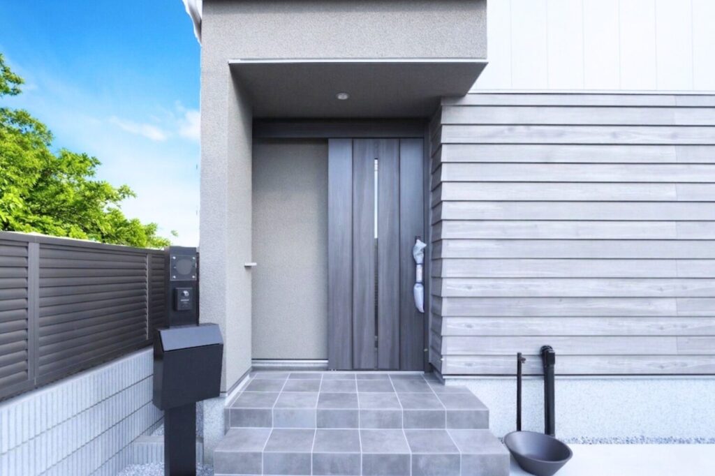 落ち着いたグレーを基調とした、モダンな一戸建て住宅の玄関アプローチ。木目調の引き戸とグレーのタイル階段、黒い機能門柱が並ぶ外観。
