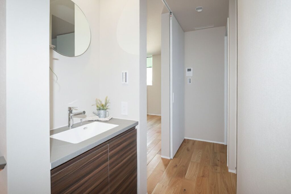 白い壁と無垢材の床の廊下に設けられたオープン洗面台。丸い鏡とグレーのカウンター、ダークブラウンの木目調キャビネットが調和する、おしゃれなナチュラルモダンの洗面スペースです。