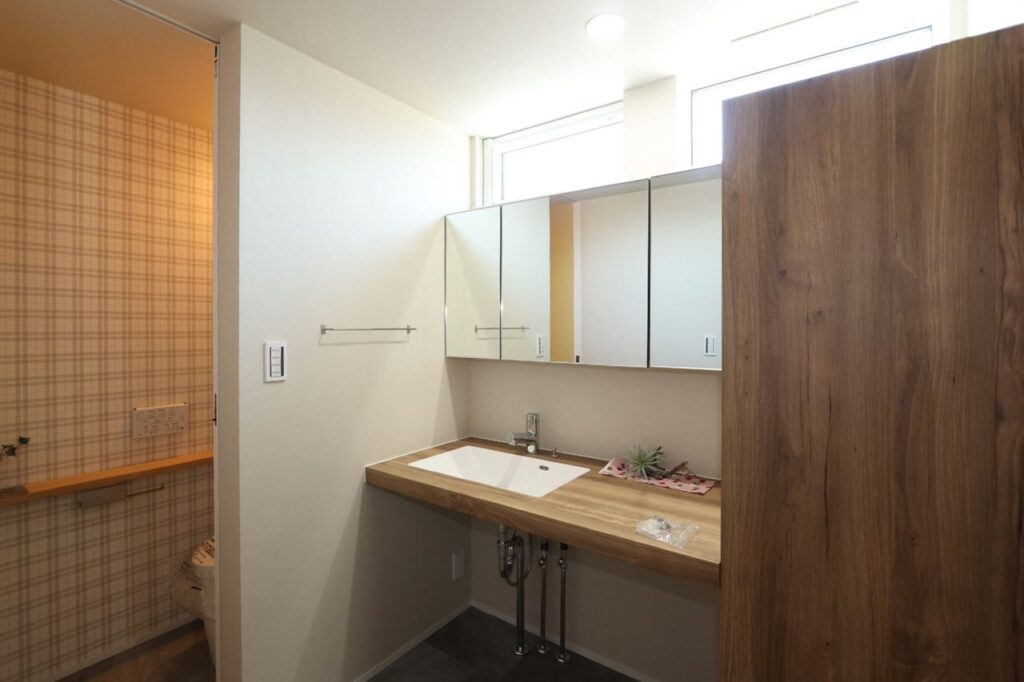 トイレに隣接するナチュラルモダンな洗面室。木目調の洗面カウンターと三面鏡、グレーの床材が調和し、鏡上部の高窓から自然光が差し込む明るく清潔感のある空間です。