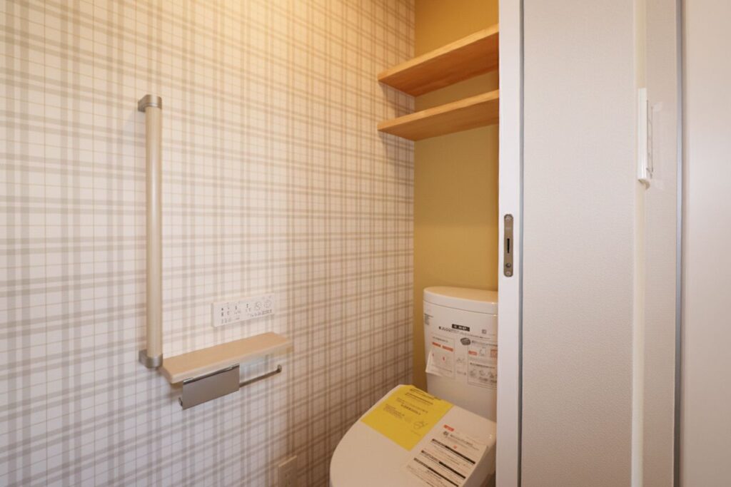 白い引き戸を開けた先にある、ベージュ系のチェック柄クロスが可愛らしいトイレ空間。立ち座りを助ける縦型の手すりや、背面上部に木製のオープン棚を備えた、温かみのある実用的なインテリアです。
