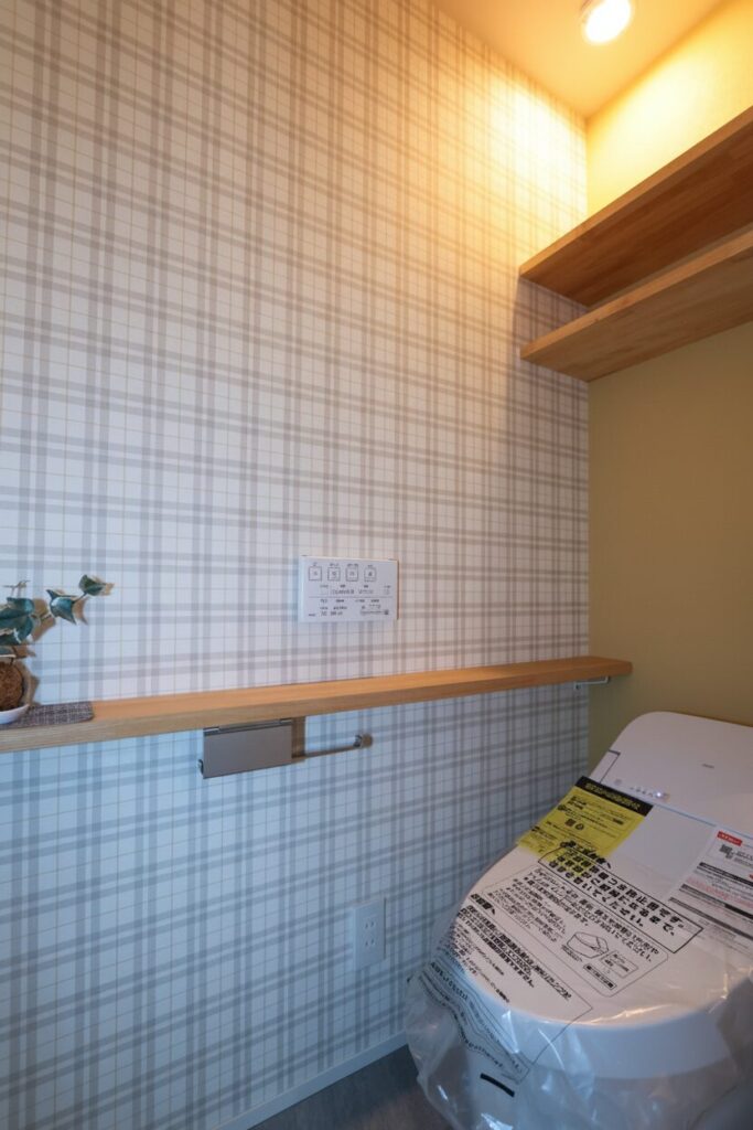 ベージュ系のチェック柄クロスが可愛らしいトイレ空間。側面の壁にはペーパーホルダーが設置された長い木製カウンターがあり、小物や植物を飾れる実用的なレイアウトです。
