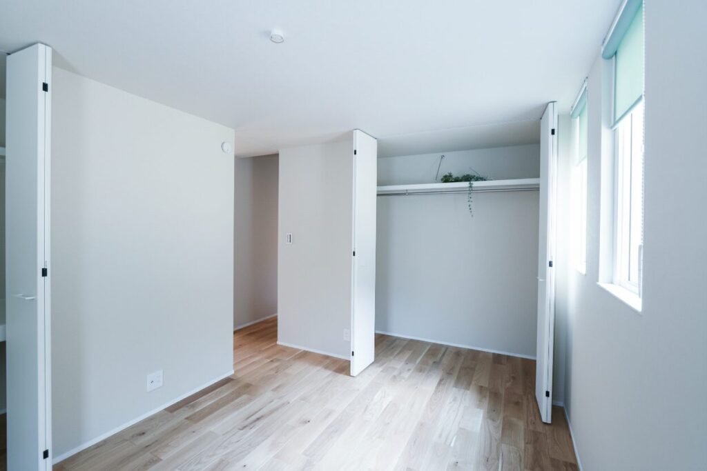 窓から明るい自然光が差し込む洋室。白い折れ戸を開けた2つのクローゼットには枕棚とハンガーパイプが備わり、無垢材の床と白い壁が爽やかなナチュラルモダンな空間です。