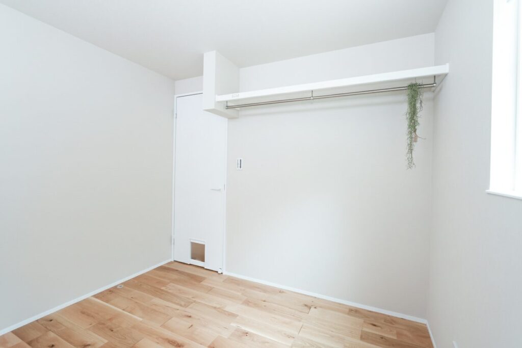 ペットドア付きの白い扉と、ハンガーパイプを備えた長い造作棚がある明るい洋室。無垢材の床と白い壁が広がる、ファミリークローゼットや室内干しスペースとして多目的に活用できるナチュラルモダンな空間です。